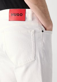 Witte denim jeans met een rood leren label met "HUGO" in reliëf, vijf-pocket ontwerp en een klassiek rechte-pijp snit.