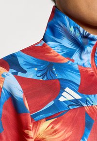 Κορυφή με ψηλό γιακά με κόκκινο και μπλε λουλουδάτο σχέδιο, υφασμένη με υφή και λευκή λεπτομέρεια λογότυπου. Διαθέτει ζωντανά μοτίβα και στενή εφαρμογή.