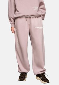 Person, der eine pinke Jogginghose mit weißem Text, einen passenden pinken Pullover und braune Sneaker trägt, steht vor einem weißen Hintergrund.
