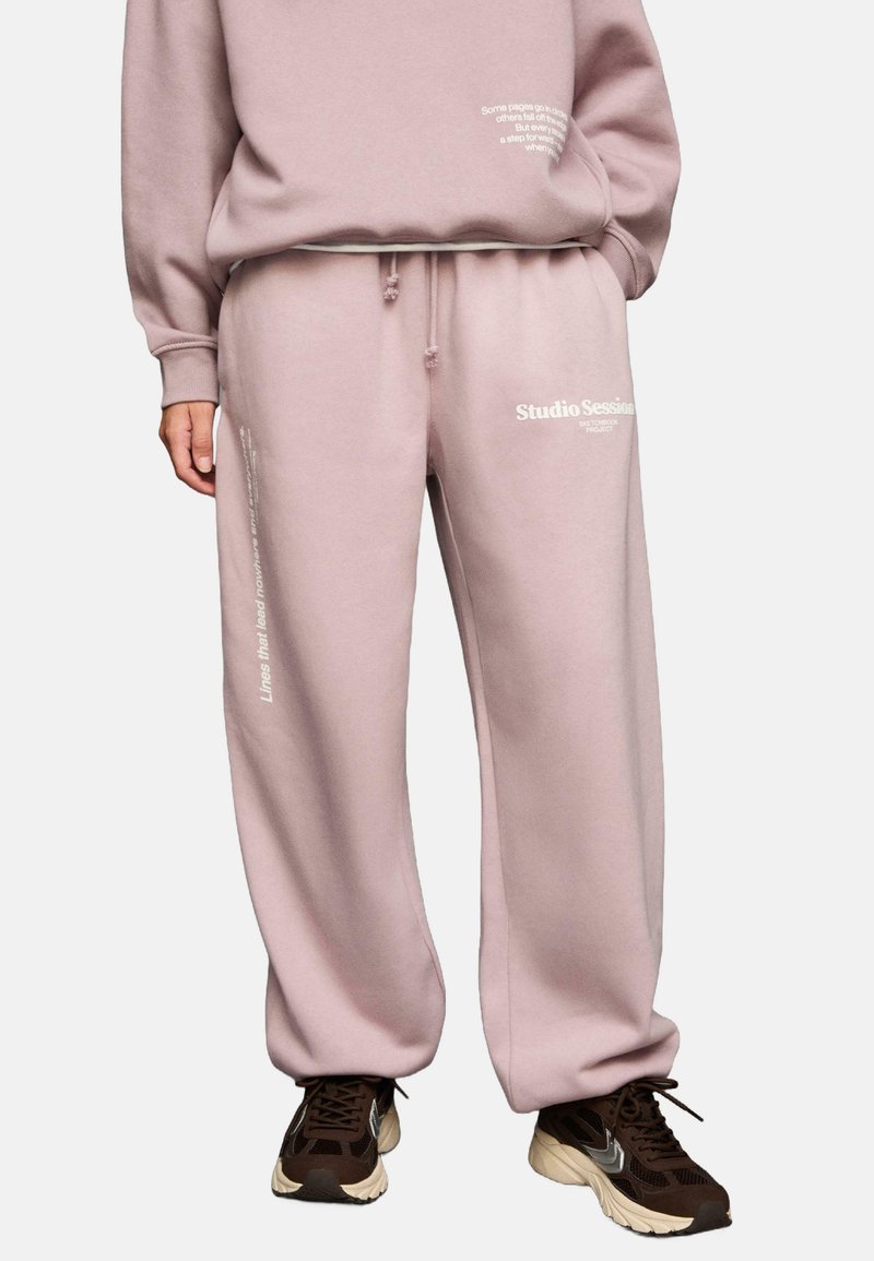 Person, der eine pinke Jogginghose mit weißem Text, einen passenden pinken Pullover und braune Sneaker trägt, steht vor einem weißen Hintergrund.