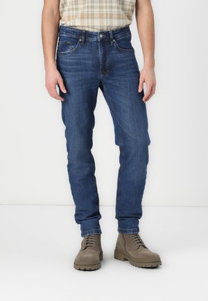 Blue denim jeans with a slim fit, featuring front pockets, a button fly, and a slight fade. Worn with beige lace-up boots.
