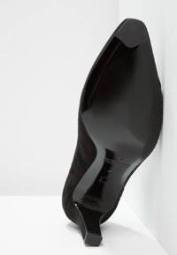 Zapato de tacón alto de ante negro con punta afilada y suela negra brillante, que presenta un tacón delgado y una textura suave.