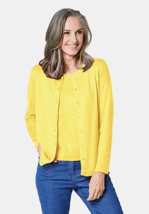 Gelber Cardigan mit rundem Halsausschnitt, Knopfleiste und langen Ärmeln, getragen über einem passenden Oberteil, kombiniert mit blauen Jeans. Glatte Textur.