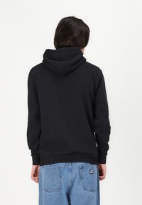 Crna dukserica s kapuljačom koja ima džep u obliku kengura, rebraste manšete i rub, u kombinaciji s svijetloplavim trapericama. Jednostavna pozadina.