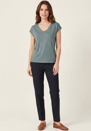 Femme debout portant un haut bleu sarcelle à manches courtes avec col en V, un pantalon slim noir et des mocassins marron sur un fond clair uni.