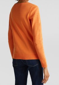 Oranger Pullover mit Rippdetails an den Ärmeln und dem Saum, aus weichem Material. Kombiniert mit dunklen Jeans.