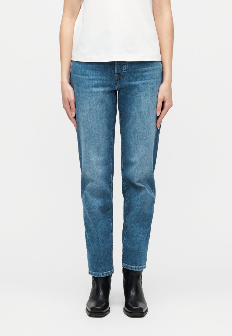 Άτομο που φορά μεσαίου ύψους μπλε τζιν και μαύρα μποτάκια στους αστραγάλους, στέκεται μπροστά σε απλό λευκό φόντο.