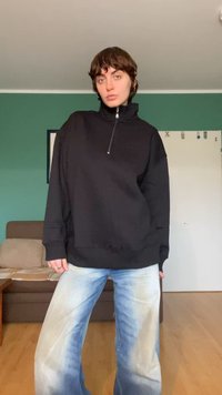 Fekete, félcipzáros pulóver enyhén texturált kivitelben, világoskék, széles szárú farmernadrággal párosítva, amelyen fakuló minták láthatók.