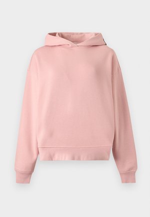 Frau trägt ein pinkfarbenes Kapuzen-Sweatshirt, lockere hellblaue Jeans und navy blaue Sneakers, steht mit einer Hand in der Tasche vor einem einfarbigen Hintergrund.