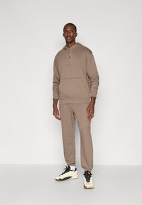 Brun hettegensere og matchende joggebukser, laget av mykt stoff. Har en frontlomme og en subtil logoaksent. Båret med hvite og svarte sneakers.