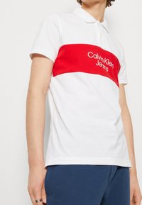 Biała koszulka polo z wyrazistym czerwonym poziomym paskiem z logiem "Calvin Klein Jeans", krótkie rękawy i klasyczny kołnierzyk.
