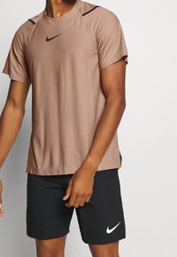 Camisa de manga corta marrón de Nike con acentos en negro en los hombros, hecha de un tejido suave; combinada con pantalones cortos negros que tienen un logo blanco de Nike.