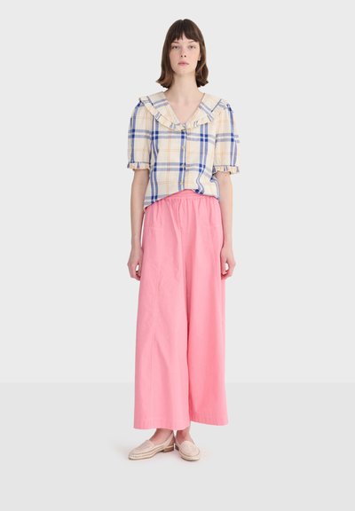 Femme portant un chemisier à manches courtes à carreaux beige et bleu avec un col à volants et un pantalon large rose, debout devant un fond uni.