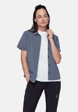 Kvinde med mørkt hår iført en kortærmet, blå ternet skjorte over en hvid t-shirt og sorte bukser, stående med den ene hånd i lommen.