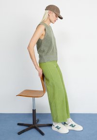 Vihreä ribbineulottu hihaton paita, vaaleanvihreät leveälahkeiset corduroy-housut, beigeillä ja vihreillä yksityiskohdilla varustetut tennarit, ruskea lippalakki ja puinen tuoli.