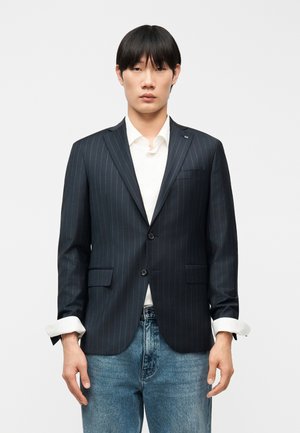 Giovane uomo che indossa un blazer a righe scure su una camicia bianca con maniche arrotolate, abbinato a jeans azzurri, in piedi contro uno sfondo neutro.