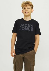 T-shirt de algodão preto com "JACK & JONES" impresso proeminentemente em cinza escuro. Mangas curtas, gola redonda. Usado com calças cargo castanhas.