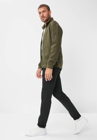 Giacca verde oliva con zip e colletto alto, tasche laterali e tessuto con texture; abbinata a pantaloni neri e sneaker bianche.