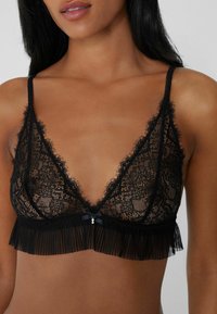 Černá krajková braletka s trojúhelníkovými košíčky, průhlednou tkaninou, volánkovým lemem a malou dekorační mašlí na přední části.