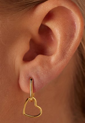 Boucle d'oreille en or en forme de cœur pendue à une oreille percée, mettant en valeur une surface métallique lisse et un design minimaliste.