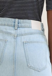 Ljusblå denimjeans med hög midja, två bakfickor och en vit etikett vid midjebandet. Tyget har en slät textur och minimala sömmar.