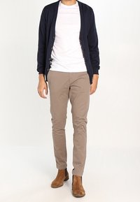 Laivastonsininen neuletakki napilla, valkoinen t-paita, vaaleanbeige housut ja ruskeat nilkkurit, seisoo vaaleataustalla.