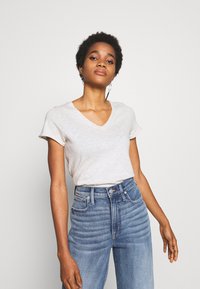 Gai pelēkas V veida kakla T-krekls ar īsām piedurknēm, apvienots ar augsta jostasvietas zilo džinsu biksēm, kas izceļas ar horizontālām šuvju detaļām un gludu tekstūru.