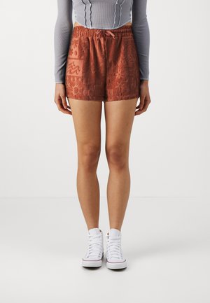 Pantalones cortos de color óxido con un patrón texturizado que presenta varias formas. Cintura elástica y detalle con cordón. Combinados con zapatillas blancas.