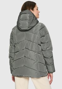 Woman wearing a gray padded jacket with a large hood, viewed from behind against a plain light background.
