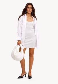 Robe mini blanche en tissu texturé avec broderie "LOVE", superposée sous une chemise blanche oversized. Tenant un sac tissé blanc. Mules noires avec accents.