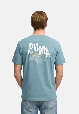 Uomo con t-shirt Puma azzurro chiaro che mostra una stampa sul retro con pallone da calcio, rete della porta, portiere e la scritta "PUMA EST. 1948".