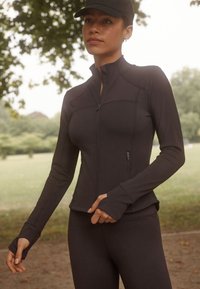 Geacă sport neagră cu guler înalt, închidere cu fermoar și manșete cu orificii pentru degetul mare. Material neted, elastic și croială slim fit.