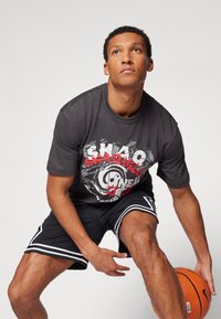 Grå bomulds t-shirt med grafisk print, parret med sorte mesh shorts med hvide striber. Modellen holder en basketball i en hinkende position.