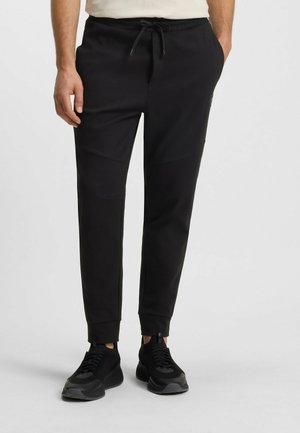 Zwarte joggers gemaakt van zacht materiaal, voorzien van een trekkoord in de taille, taps toelopende pijpen en gedetailleerde panelenstiksels. Draagt zwarte sneakers.