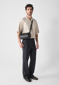 Bolso cruzado de cuero negro con forma redondeada, llevado sobre una camisa de manga corta beige y pantalones azul marino, con zapatos texturizados.