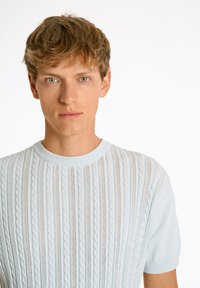 Ljusblå stickad tröja med kabelmönster och korta ärmar, med en ribbad halsringning. Tyget har en texturerad, ventilerande kvalitet.