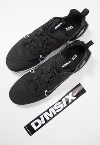 Black athletic shoes made of mesh with embossed texture, featuring white Swoosh logo and branding, accompanied by a black promotional sticker.