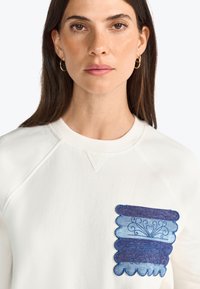 Felpa bianca con una particolare tasca in denim a gradiente blu, caratterizzata da bordi scallop e motivi di cucitura decorativa.