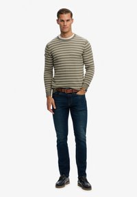 Suéter a rayas en verde oliva y crema con cuello redondo, combinado con jeans azul oscuro y zapatos negros. Cinturón de cuero marrón y reloj visibles.