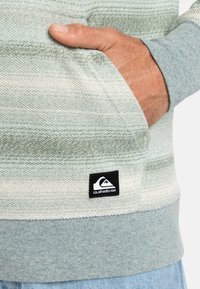 Suéter a rayas verde claro y blanco con una textura suave, que presenta un pequeño logo negro de Quiksilver bordado en el bolsillo.