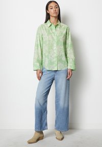 Blusa verde con motivo floreale, maniche lunghe e chiusura a bottoni, abbinata a jeans a gamba larga blu chiaro e stivaletti beige.