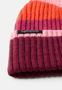 Bonnet en tricot avec un motif rayé dans des tons d'orange, de rose et de bordeaux. Accentué par une étiquette noire portant la mention "PeakPerformance."