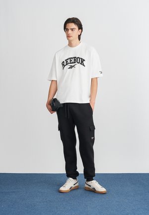Hvid oversized t-shirt med sort Reebok-logo, sorte cargo-joggers og cremefarvede sneakers med sorte og gummi såler. Simpelt og sporty design.