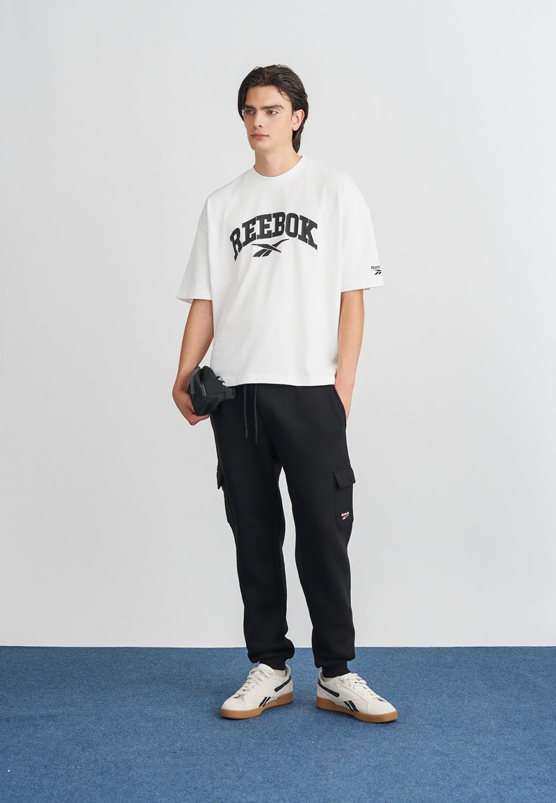 Bela oversized majica s črnim Reebok logotipom, črni cargo joggerji in smetanasto beži superge s črnimi in gumijastimi podplati. Enostaven in športen dizajn.
