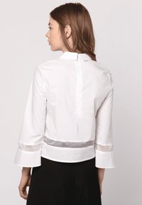 Camisa blanca con mangas de campana 3/4, cuello y apertura en la parte posterior. Presenta una banda transparente horizontal en la cintura y los dobladillos de las mangas. Textura suave.