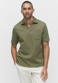 Lyhythihainen oliivinvihreä polo-paita, jossa on v-aukko kaulus, texturoitu kangas ja rintatasku. Yhdistetty vaaleisiin housuihin.