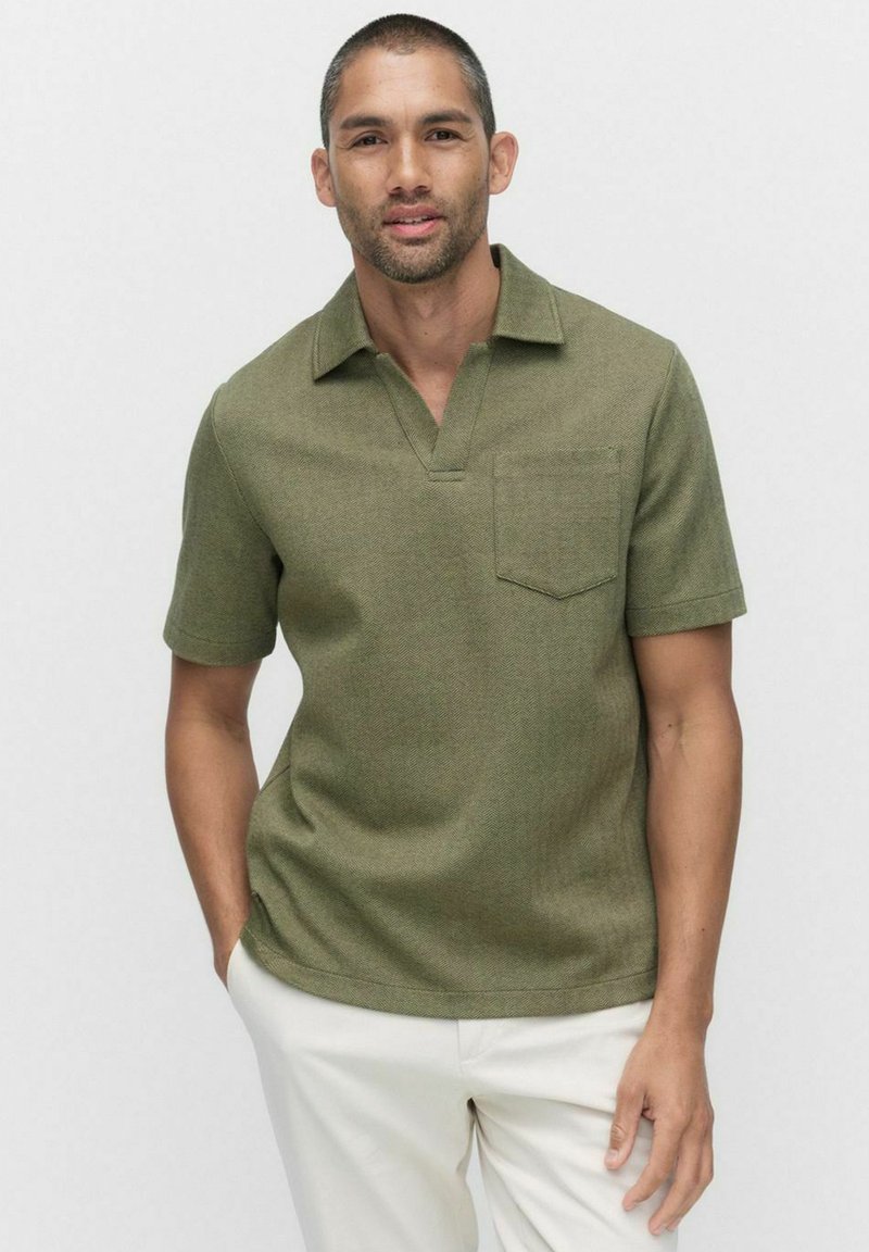 Lyhythihainen oliivinvihreä polo-paita, jossa on v-aukko kaulus, texturoitu kangas ja rintatasku. Yhdistetty vaaleisiin housuihin.