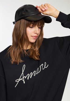 Cappello di pelle nera con una visiera leggermente curvata, indossato da una persona in felpa nera con testo in corsivo bianco "Amore." Texture liscia.