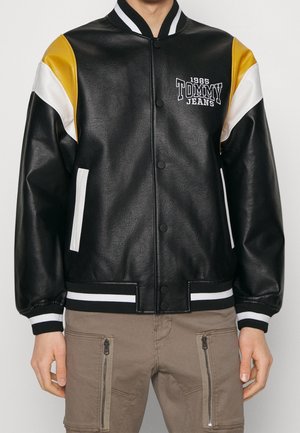 Mand iført sort læder varsityjakke med hvide og gule skulderaccents, hvide stribede manchetter og "1985 Tommy Jeans" logo.