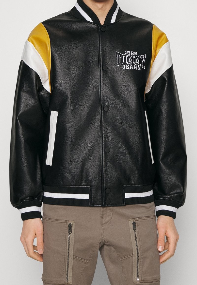 Homme portant une veste varsity en cuir noire avec des accents blancs et jaunes aux épaules, des poignets blancs rayés et le logo « 1985 Tommy Jeans ».